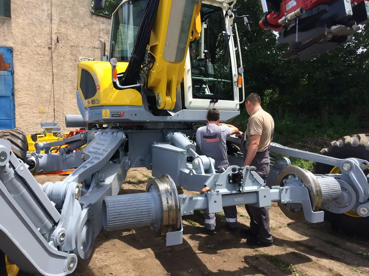 Mecanici montând sistemul de deplasare pe șine pe un Menzi Muck M545X înainte de predarea la CFR Brașov