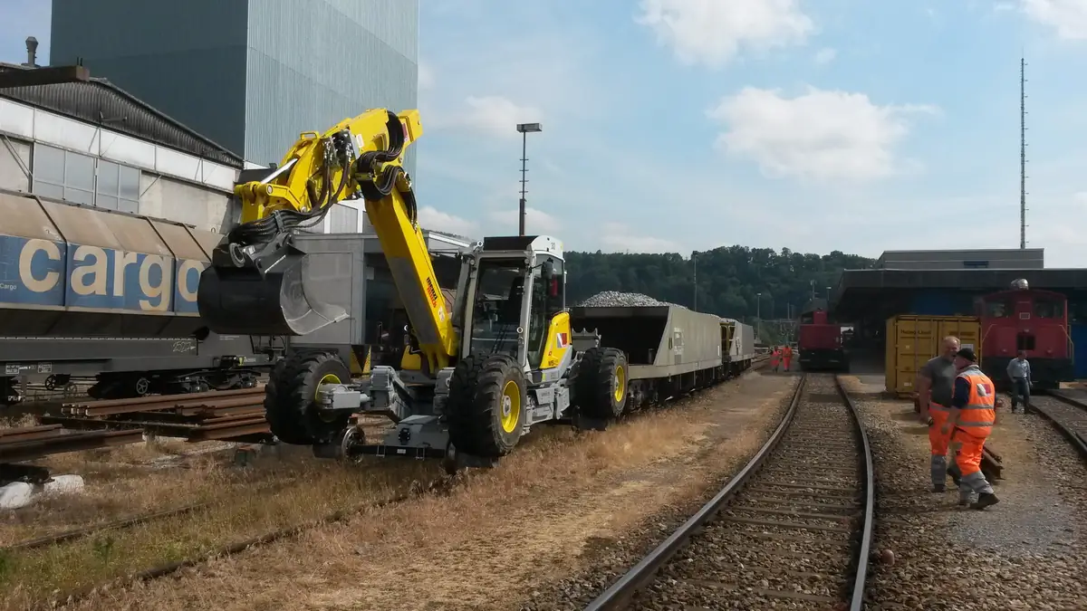 Menzi Muck M545 pe roți de cale ferată, cu cupă, într-o stație de marfă