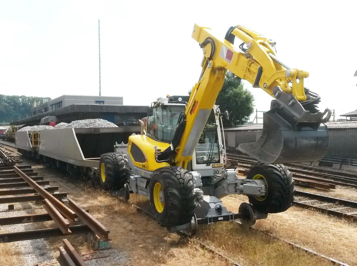 Roțile de cale ferată ale unui Menzi Muck M545, lângă vagoane de balast