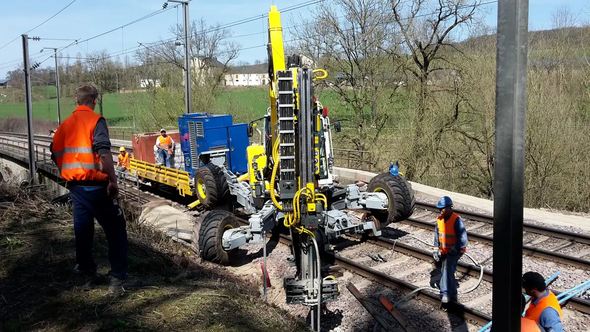 Menzi Muck M545 cu foreză pe braț, cu picioarele de o parte și de alta a liniei, între muncitori