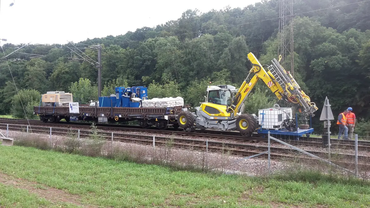 Menzi Muck M545 cu foreză, pe șine, tractând un vagon platformă cu compresor
