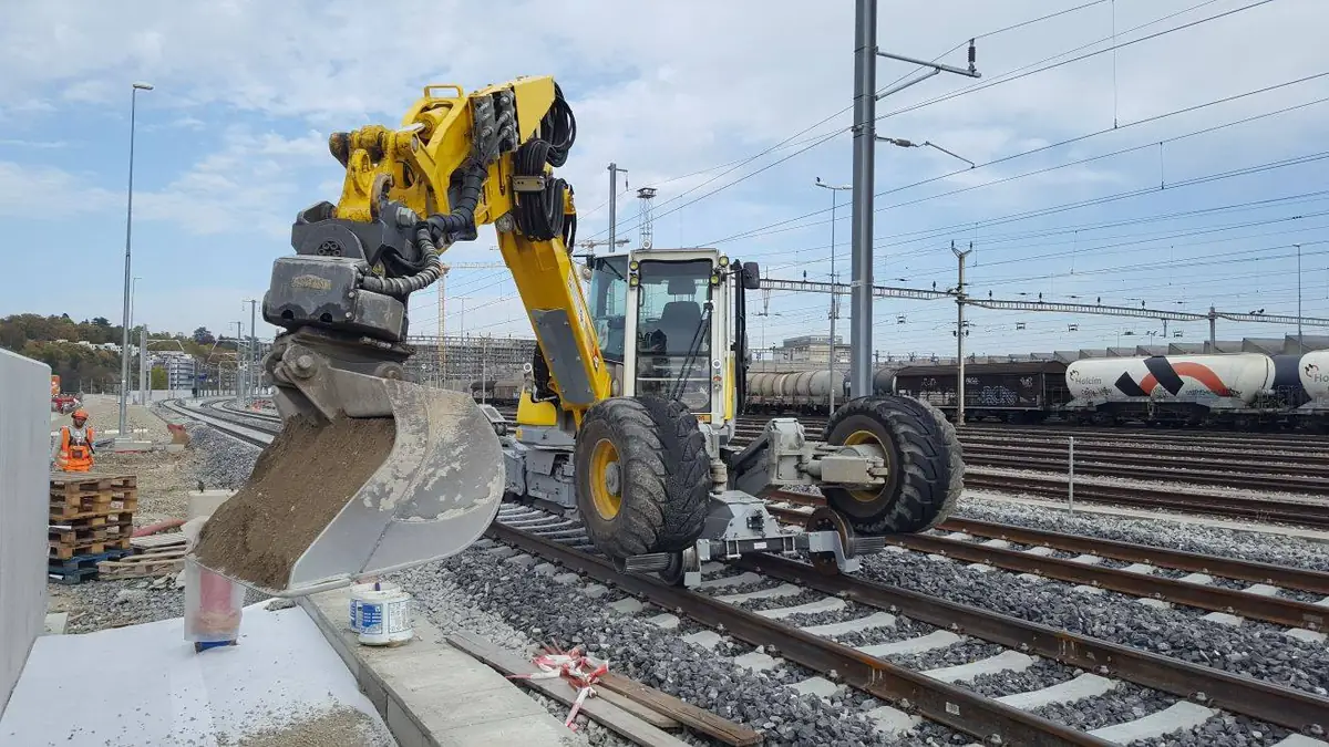 Menzi Muck M545 pe șine descărcând pământ cu cupa pe un șantier de stație