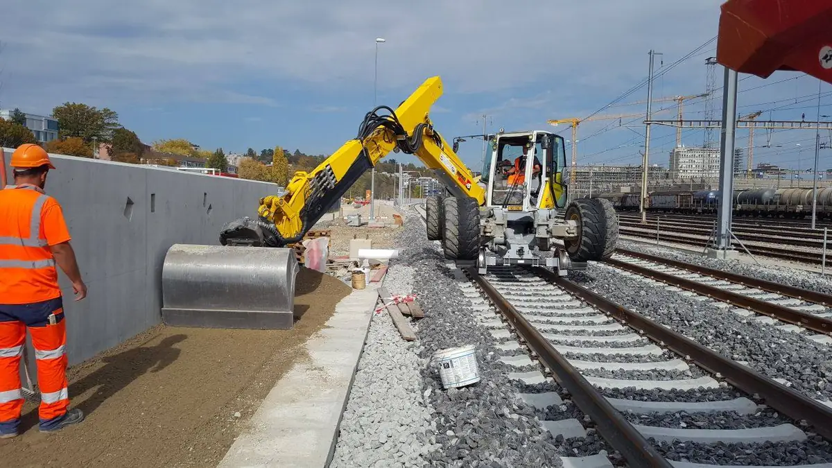 Excavator pe picioare Menzi Muck M545 pe șine, cu cupa lângă un zid de beton, din arhiva Baier Solutions