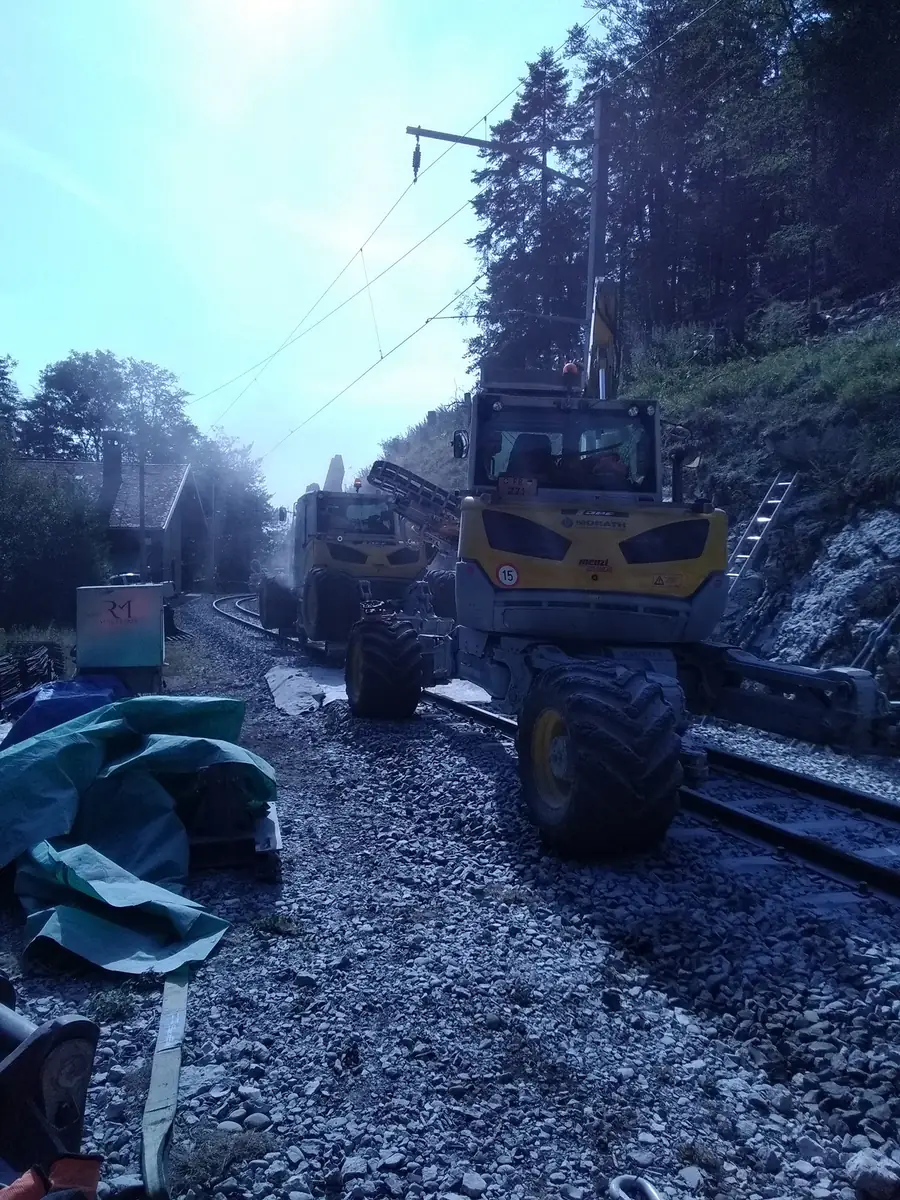 Două Menzi Muck M545 pe o linie ferată în curbă, văzute din spate, în timpul forării la taluz