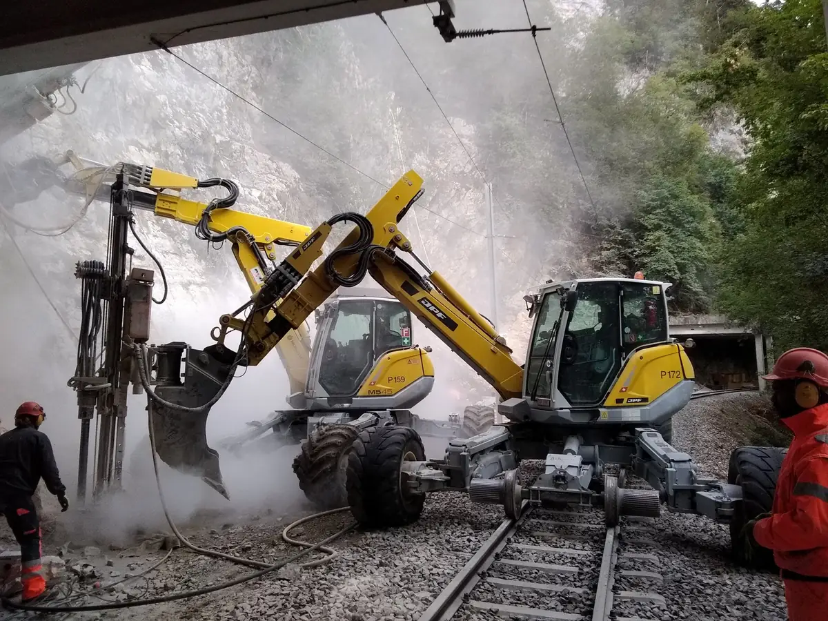 Două Menzi Muck M545 sub o galerie de protecție a căii ferate: unul forează în stâncă, celălalt stă pe roțile de cale ferată