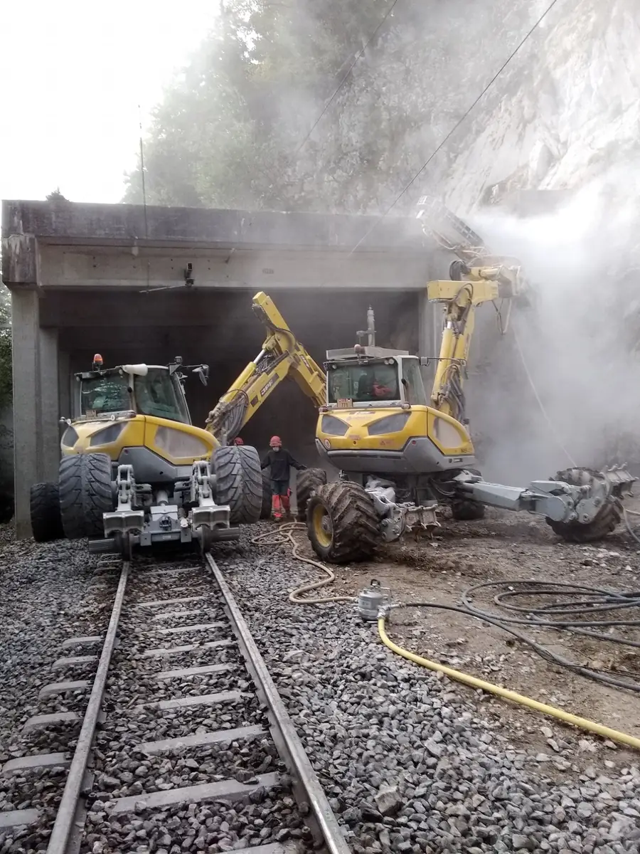 Două excavatoare pășitoare la intrarea unei galerii de cale ferată, unul pe șine și unul cu picioarele pe terasament